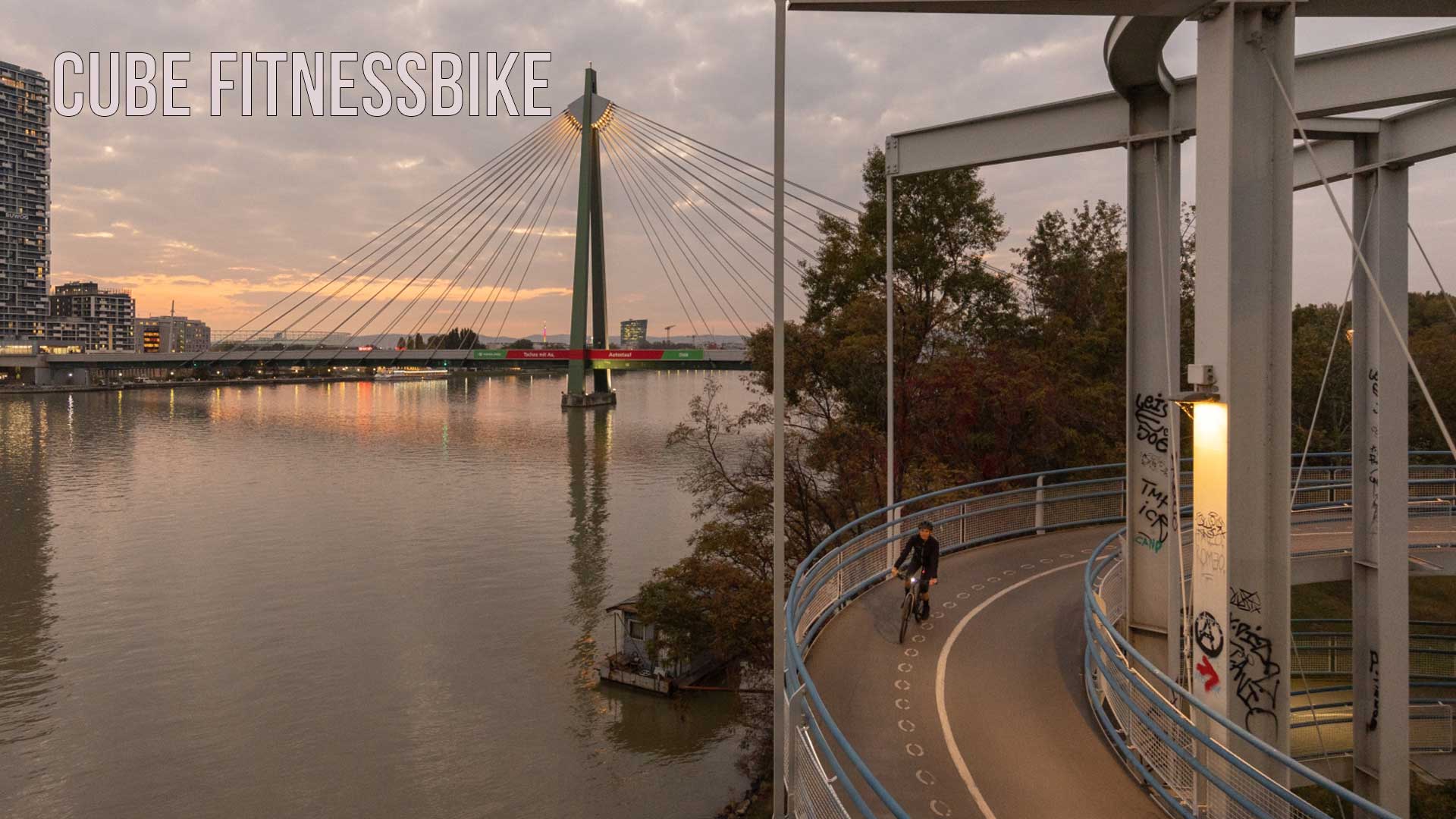Man ser en by ved floden. I forgrunden ses en cyklist, der kører på cykelstien i skumringen. Billedet er forsynet med titlen »Cube Fitnessbike«.