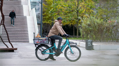 Man ser en mand på en blå Cube Longtail Hybrid-lastcykel, der kører gennem bymidten med dagligvarer på cyklen.