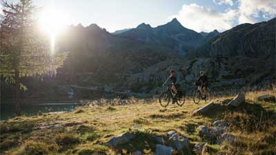Et par cykler på deres trekkingcykler ad en vandresti. I baggrunden ses bjerge, bag hvilke solen titter frem.