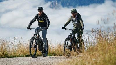 To cyklister kører på deres el-mountainbikes på en cykelsti. I baggrunden ses bjerge.