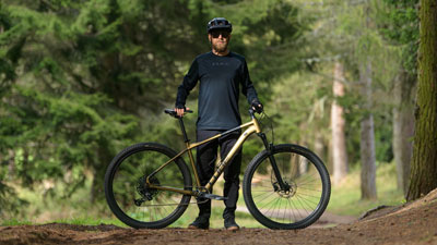Man ser en mand bag sin gyldne Cube-mountainbike på en skovvej.