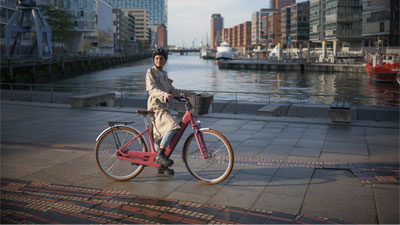 Man ser en kvinde på en lyserød Cube Ella Hybrid-elcykel. Hun cykler langs en flod. I baggrunden ses en by.