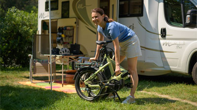 Man ser en kvinde, der står foran en autocamper. Foran hende står en sammenklappet grøn foldecykel.