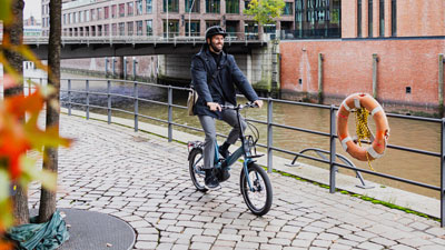 En mand kører på sin blå foldecykel; i baggrunden er huse og en flod.