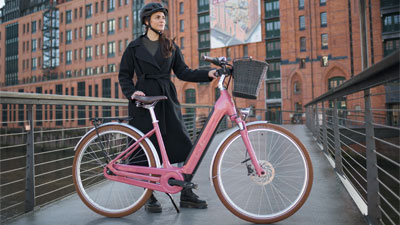 På billedet ses en kvinde bag en lyserød Cube Ella Hybrid-elcykel. I baggrunden ses en bygning.