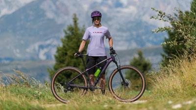 Man ser en kvinde i lilla cykeltøj, og foran hende står en lilla Cube-mountainbike. I baggrunden ses et bjergrigt landskab.