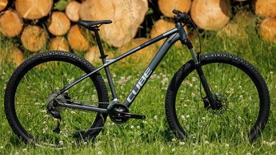 Man ser en grå Cube-mountainbike, der står på en eng. I baggrunden er der stablet brænde.