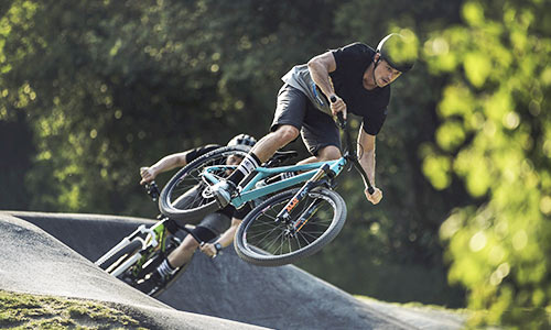 En cyklist hopper på en turkisblå dirtbike i en cykelpark; i baggrunden ses en anden cyklist på en grøn cykel