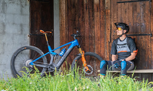 Man ser en cyklist, der sidder på en bænk foran en lade. Ved siden af ham står hans fuldt affjedrede e-mountainbike