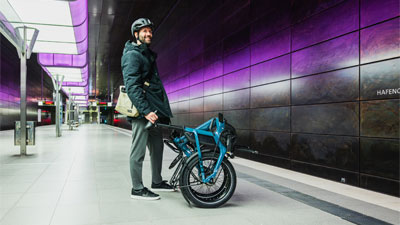Man ser en mand, der står på en metrostation. Foran ham står en sammenklappet blå foldecykel.