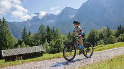 En dreng cykler på sin mountainbike ad en grusvej gennem et bjergrigt landskab. I baggrunden ses en hytte.