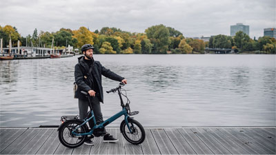 En mand står ved siden af sin blå sammenklappelcykel på en bro. I baggrunden ses vandet og en by.