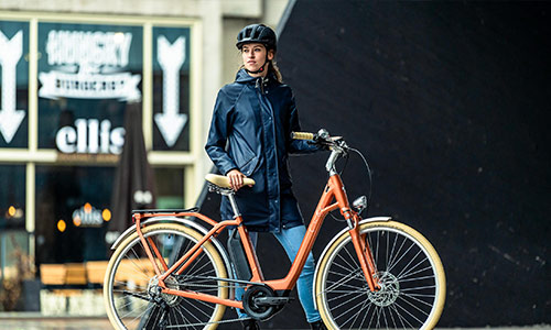 En kvinde står bag sin orange bycykel i bymidten.
